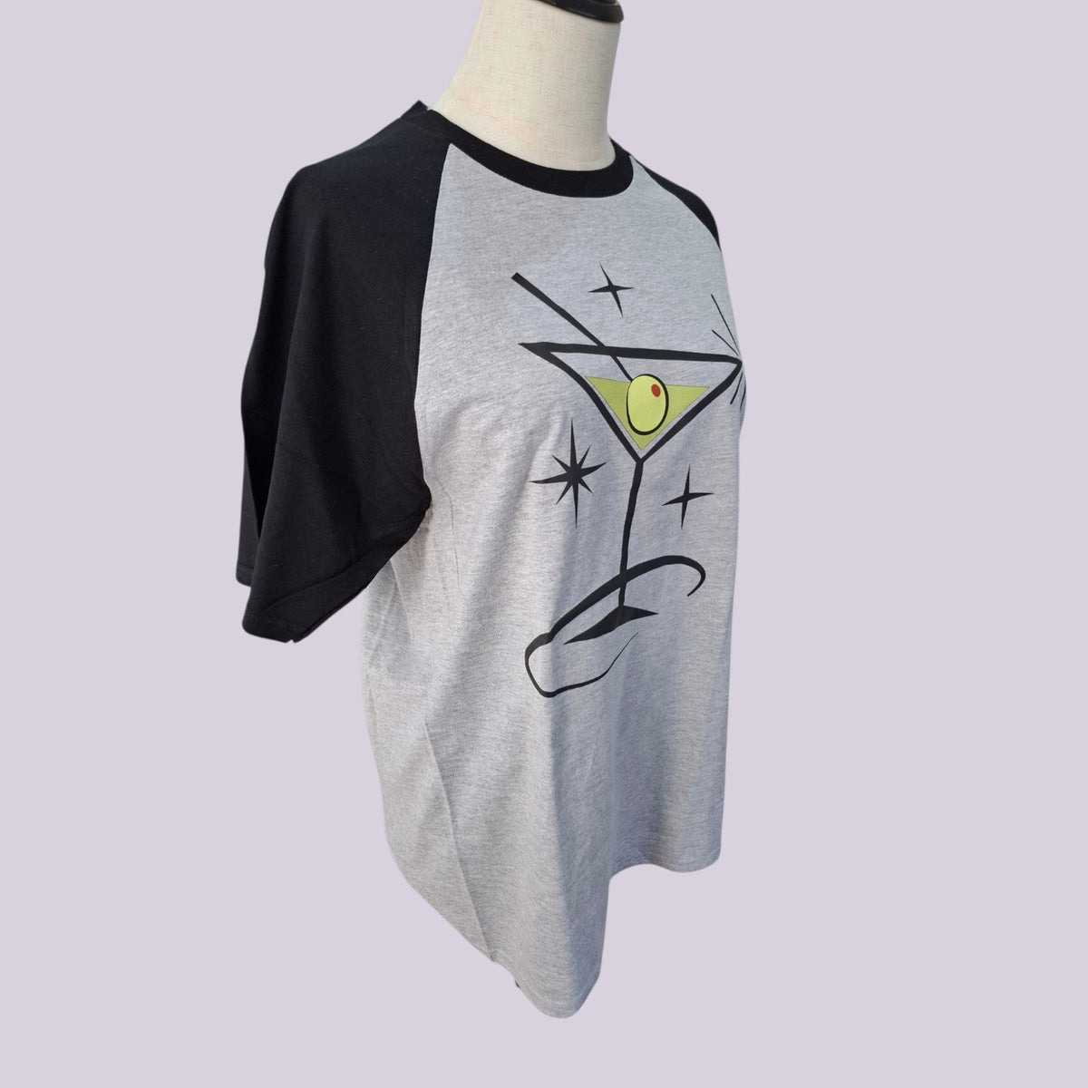 A heather gray and black raglan t-shirt displayed on a mannequin torso. The shirt features a graphic of a martini glass with a green olivine and black sparkles on the front, set against a solid light purple background.