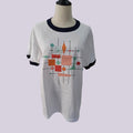 A white ringer t-shirt with a colorful mid-century modern geometric print on the front, displayed on a black mannequin torso against a light lavender background.