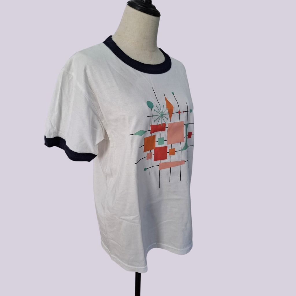 A white ringer t-shirt with black trim on the collar and sleeve cuffs, featuring a colorful abstract geometric print on the front, displayed on a mannequin torso against a light lavender background.