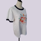 A white ringer t-shirt with black trim on the collar and sleeve cuffs, featuring a colorful abstract geometric print on the front, displayed on a mannequin torso against a light lavender background.