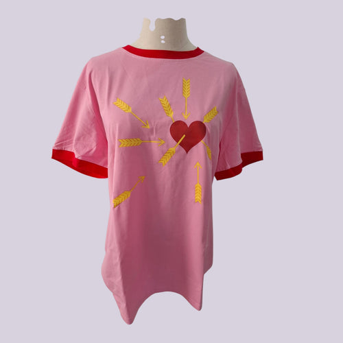 A pink short-sleeve ringer t-shirt displayed on a mannequin torso. The shirt features a red crew neck and sleeve cuffs, with a graphic print of a red heart pierced by yellow arrows and radiating yellow wheat stalks.
