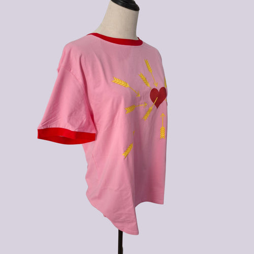 A pink short-sleeve ringer t-shirt with red trim on the collar and sleeve cuffs, featuring a graphic of a red heart and yellow wheat stalks on the front, displayed on a mannequin torso against a light gray background.