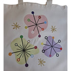 A white canvas tote bag featuring a retro mid-century modern design with three colorful abstract shapes, starburst patterns, and star-like accents in pink, purple, green, yellow, and gold.