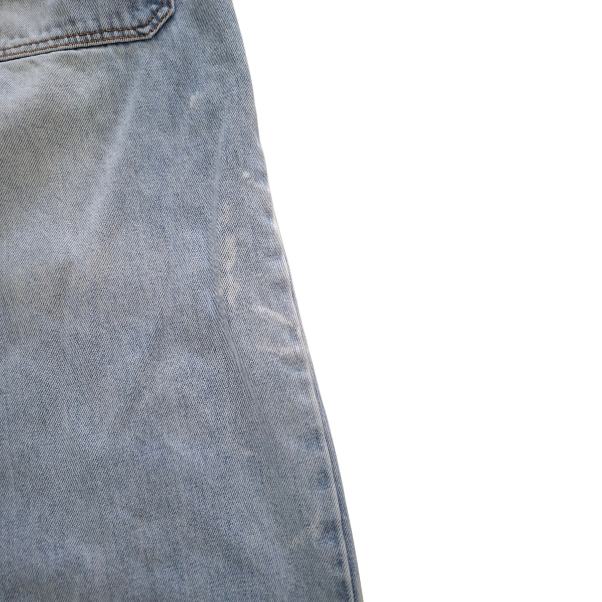Close-up of a light blue denim fabric, showing the texture and a portion of a pocket seam against a plain white background.