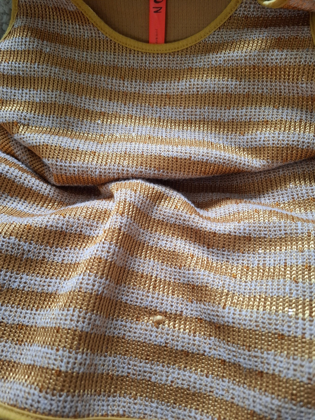 Close-up of a striped knit garment in warm mustard yellow and off-white, showing the textured fabric and part of a bright orange neck label.