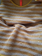 Close-up of a striped knit garment in warm mustard yellow and off-white, showing the textured fabric and part of a bright orange neck label.