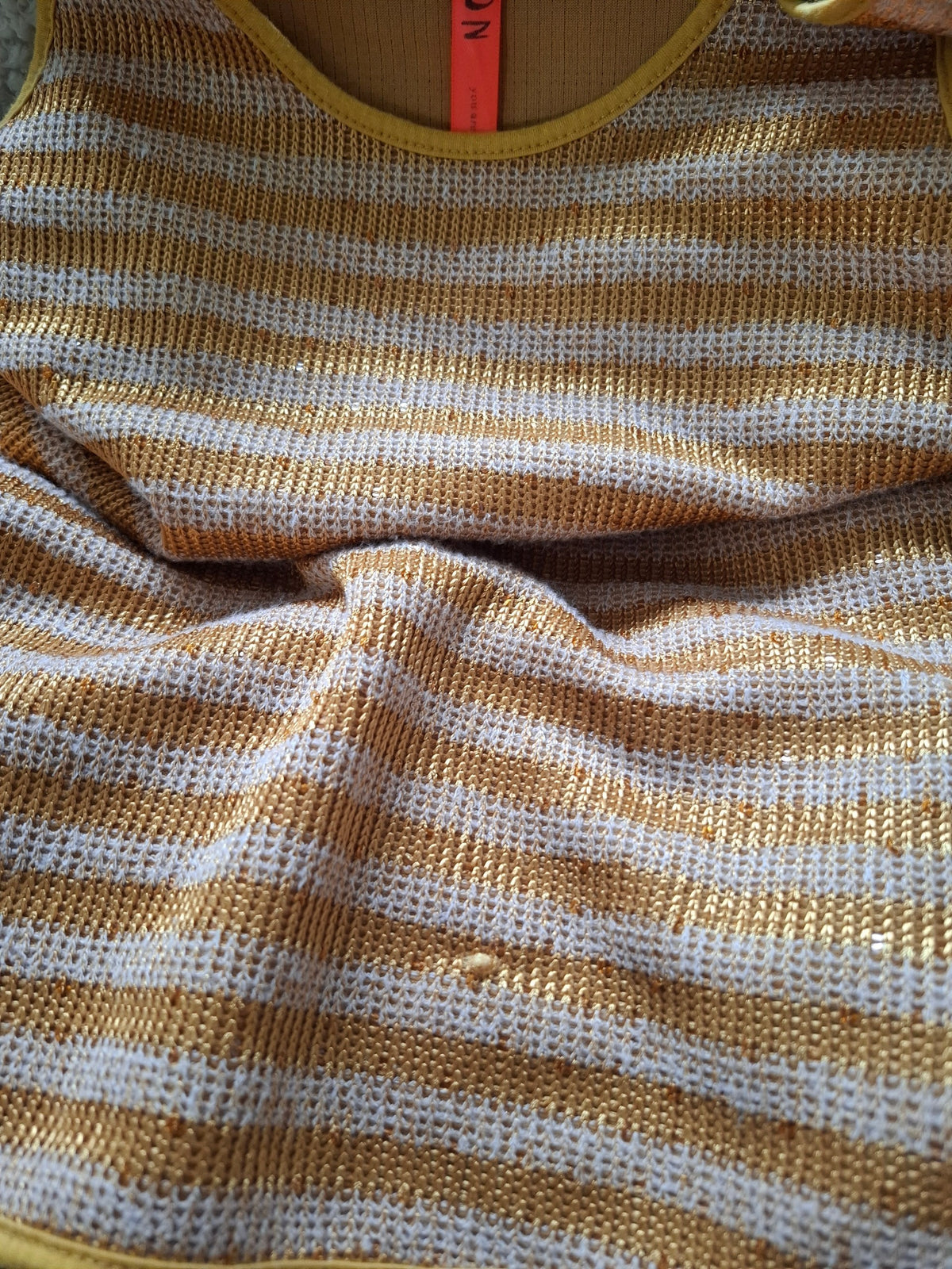 Close-up of a striped knit garment in warm mustard yellow and off-white, showing the textured fabric and part of a bright orange neck label.
