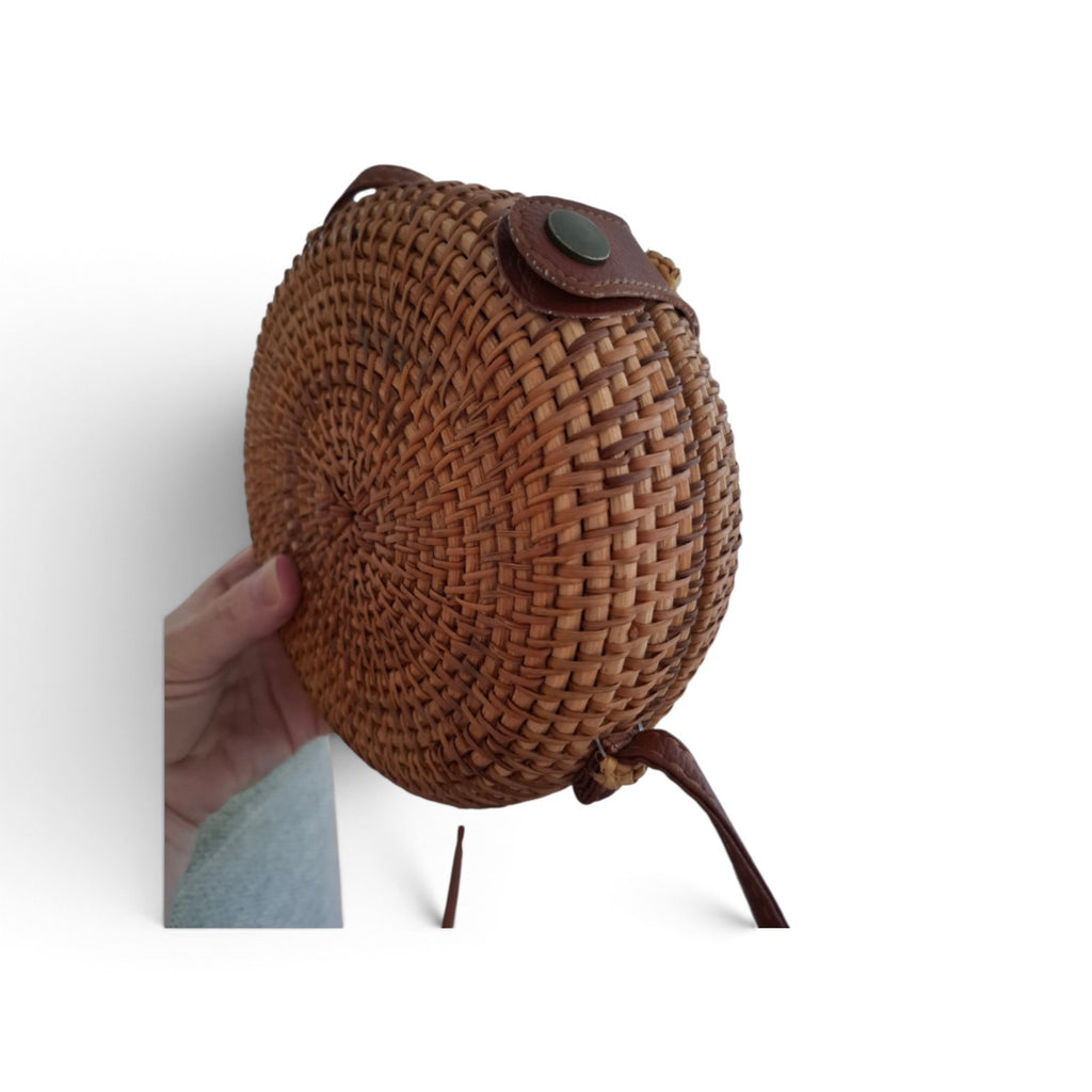 A round woven straw bag with dark brown leather trim and a small green button closure, held by a hand against a white background.