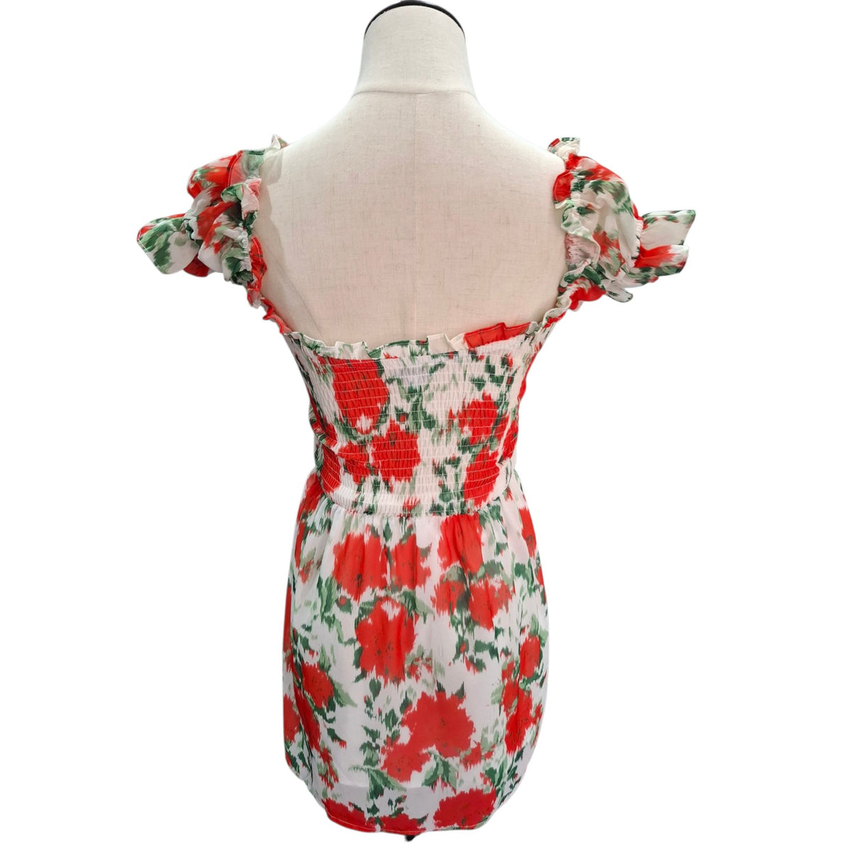 A white smocked mini dress with a red and green floral print, displayed on a cream-colored dress form mannequin against a white background.