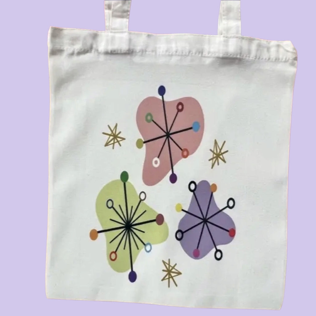 A white canvas tote bag with two handles, featuring a retro mid-century modern design with three abstract organic shapes in pink, green, and purple, accented with black radial lines and small colored dots, set against a solid lavender background.