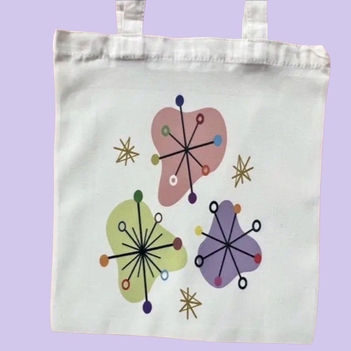 A white canvas tote bag with two handles, featuring a retro mid-century modern design with three abstract organic shapes in pink, green, and purple, accented with black radial lines and small colored dots, set against a solid lavender background.