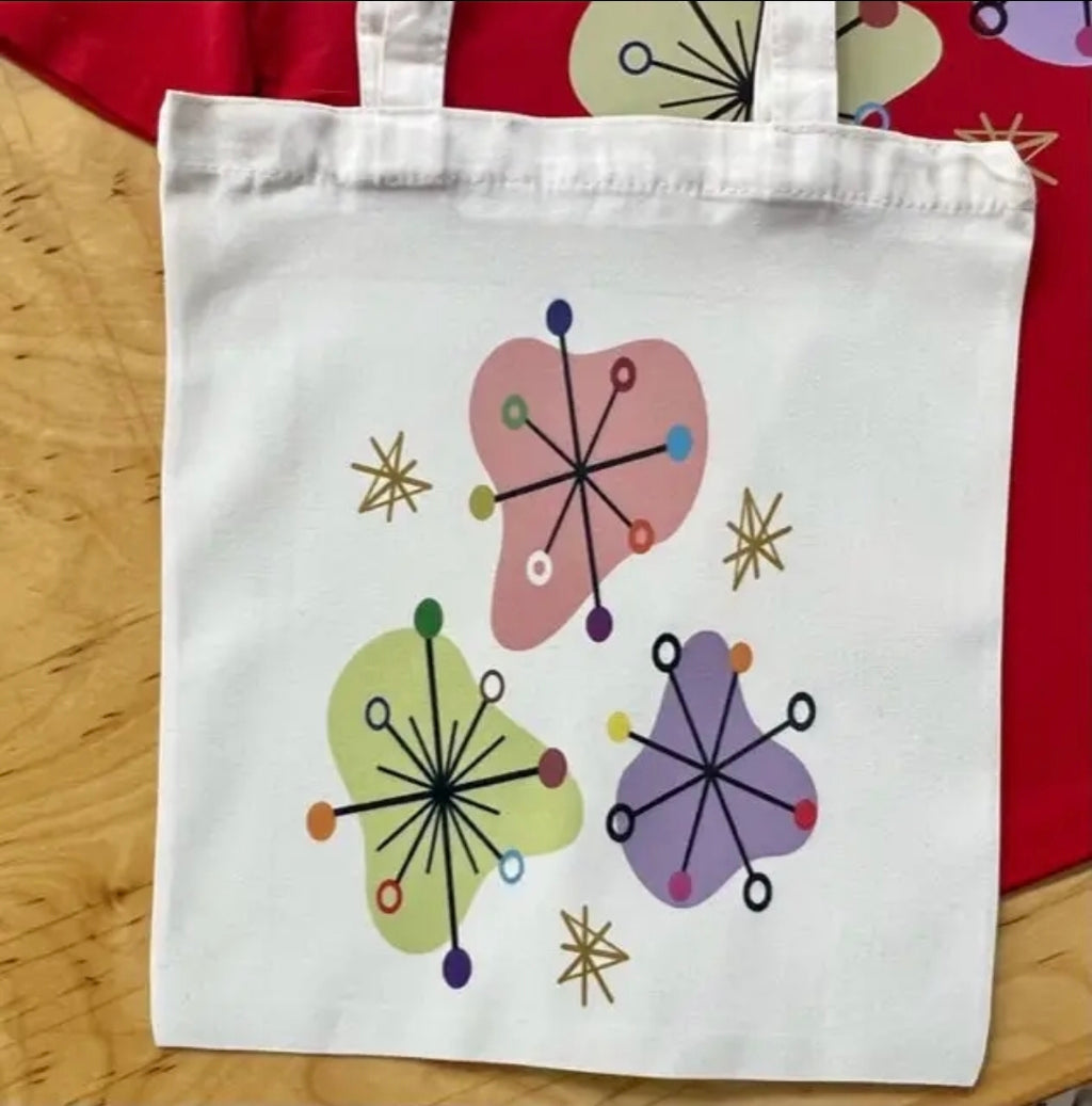 A white canvas tote bag with a colorful mid-century modern abstract floral design, displayed on a light wood surface with a red fabric background.