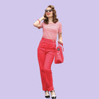 A woman modeling a red and white striped short-sleeve top, bright red pants, black and white heels, sunglasses, and a red handbag against a solid light purple background.